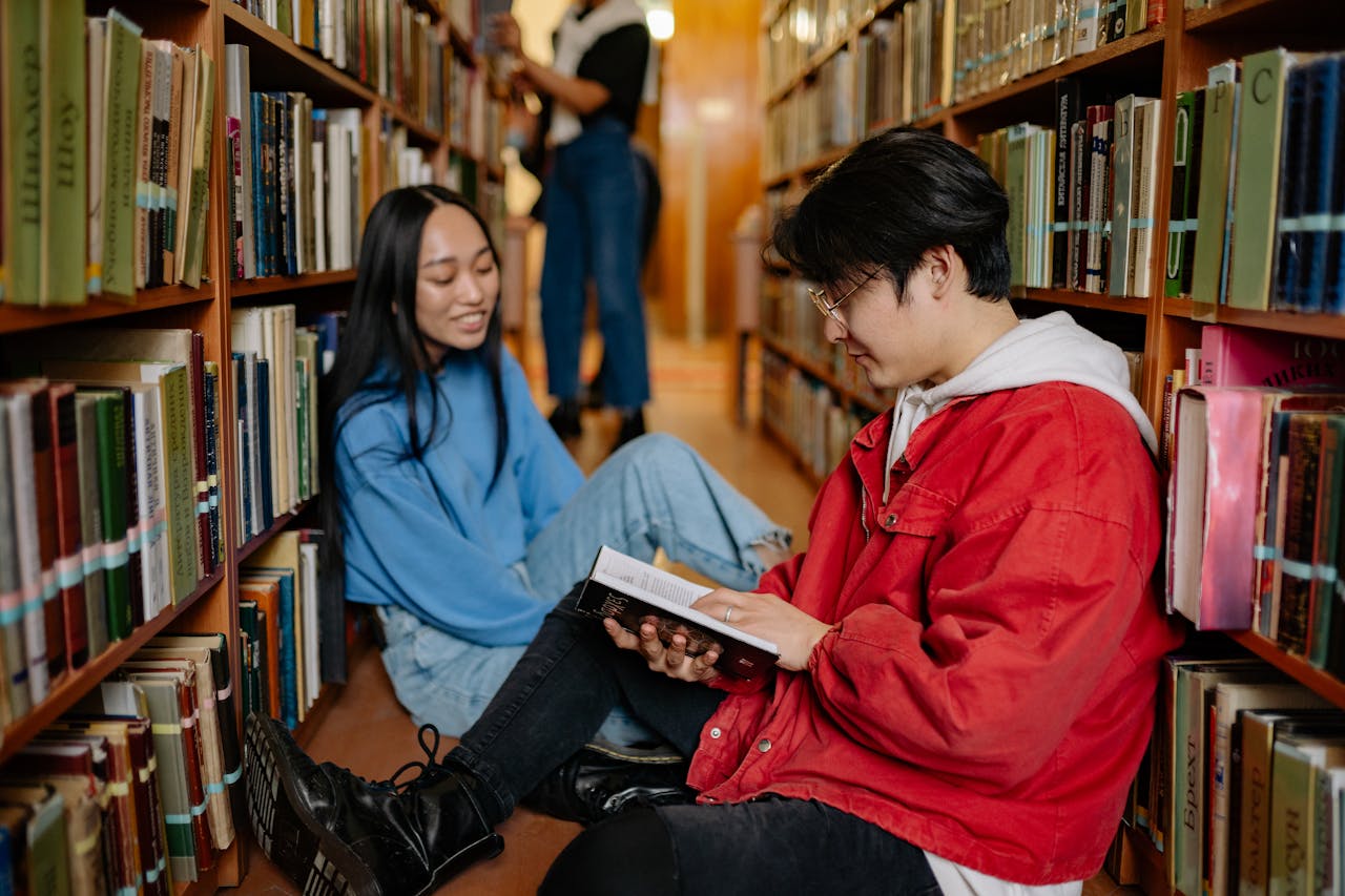 Two students sitting on a library floor reading and discussing books.